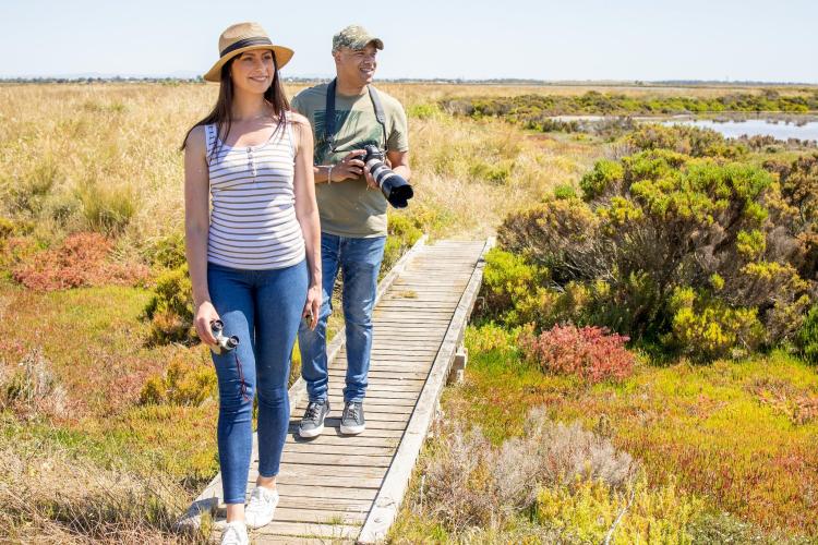 two adults with large camera walking along path in wetlands