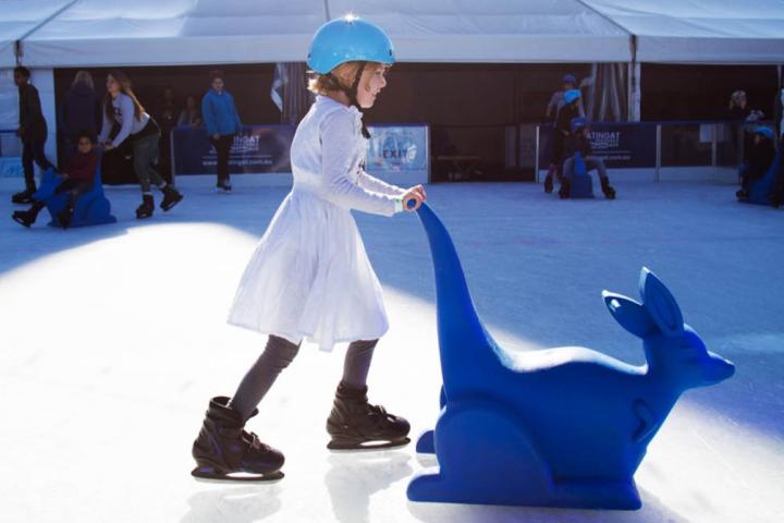 child in a white dress ice skating using a kangaroo skating aid
